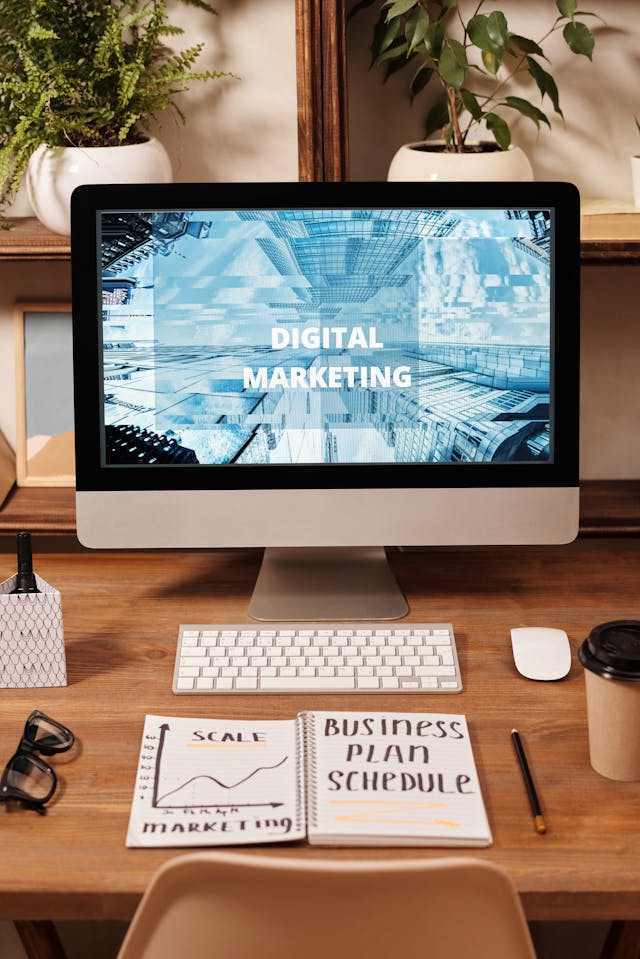 Desktop computer displaying “Digital Marketing” on a tidy wooden desk with notebook, keyboard, mouse, and coffee cup